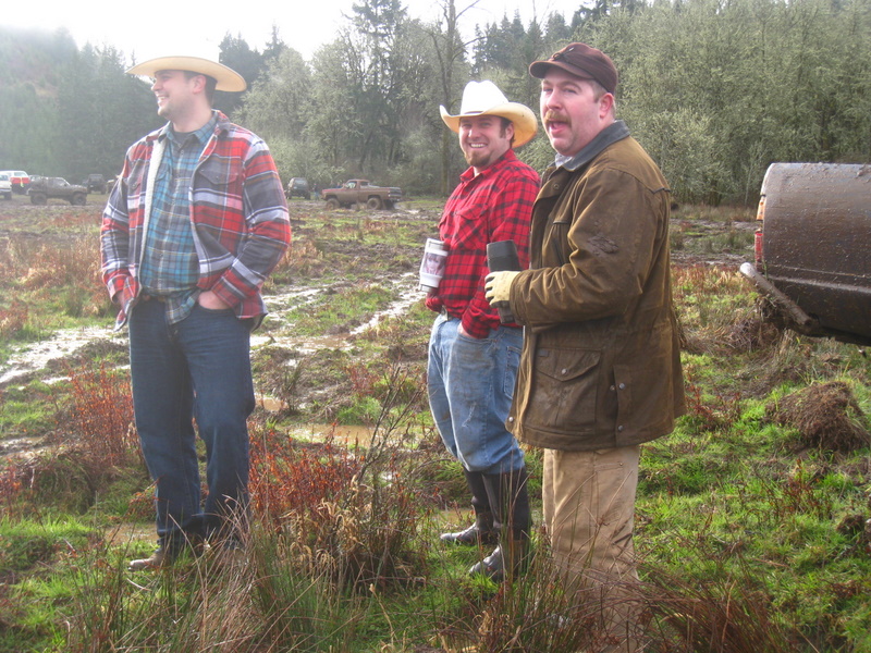 Three men in cowboy hats and flannel shirts stand in a muddy field. Trucks are visible in the background