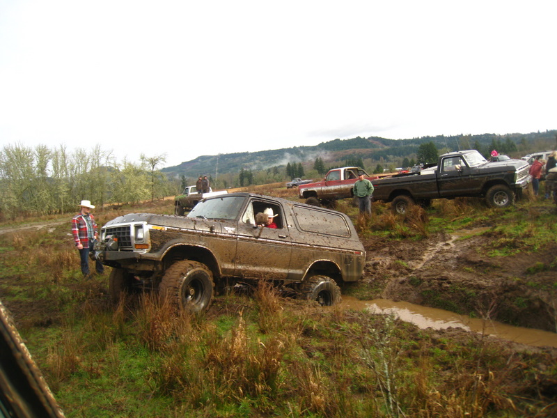 Mud-covered 1978 Ford Bronco with a person in a cowboy hat driving, in a muddy field with other trucks