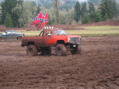 1978 Ford Bronco - A lifted red pickup truck navigates a muddy field, with two confederate flags prominently displayed.