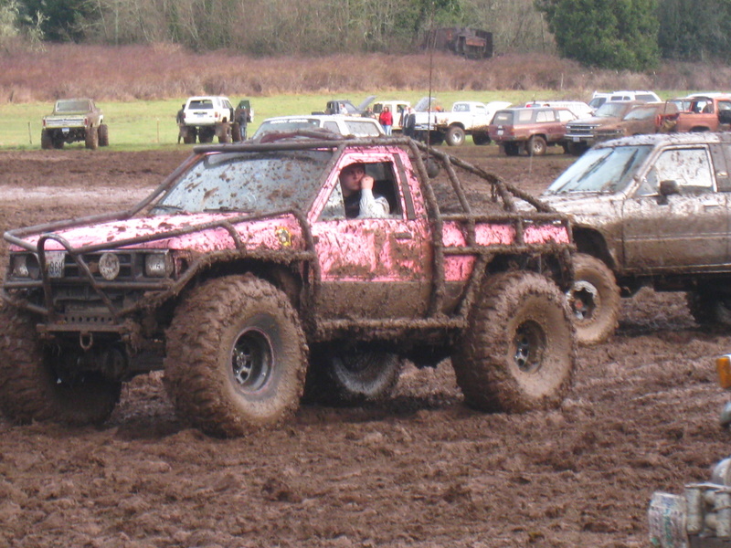 Pink pickup truck with roll cage and large tires in mud. Driver visible