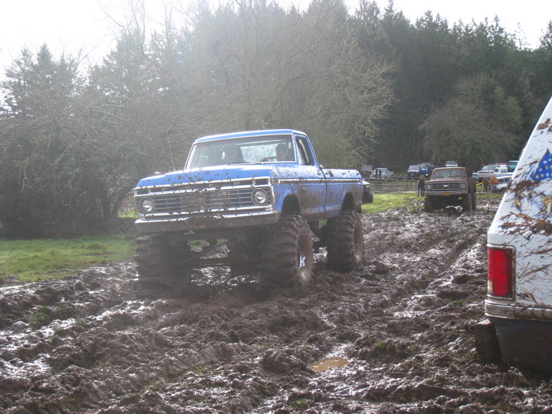 Blue lifted pickup truck with large tires driving through mud. Another truck is visible in the background