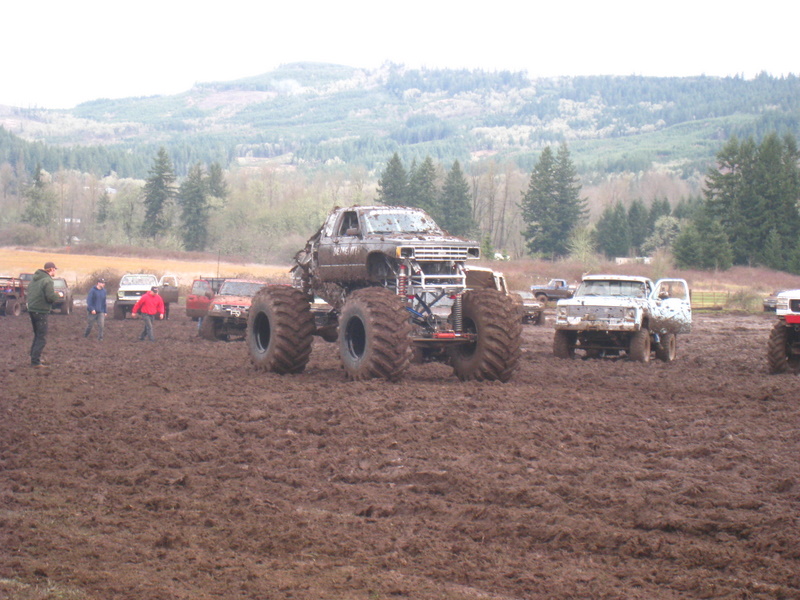 Monster truck with large tires sits in mud, other trucks and people in background