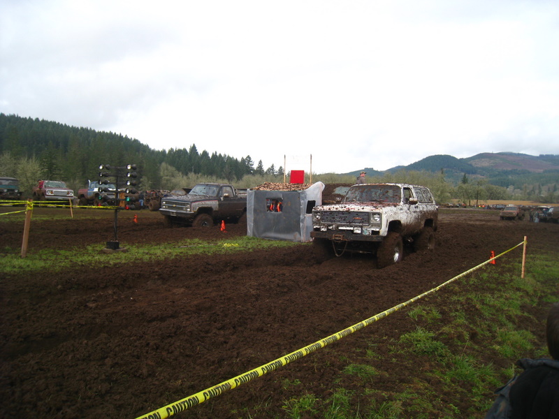 White SUV stuck in mud, front view. Other vehicles and muddy terrain visible