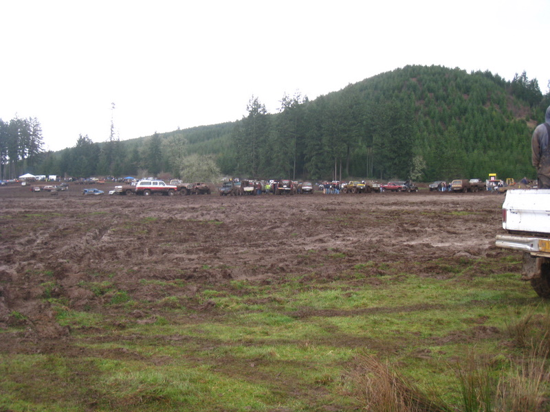 Muddy field with many trucks and SUVs parked in the distance. A person stands on the back of a white truck in the foreground