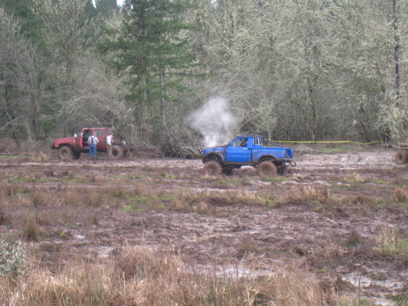 Blue pickup truck emitting steam in a muddy field with a red truck nearby