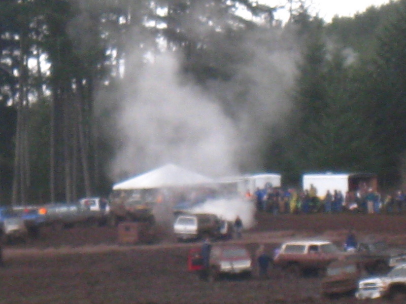 Mud bog event with several vehicles, tents, and a crowd of spectators. Smoke or steam rises from one area