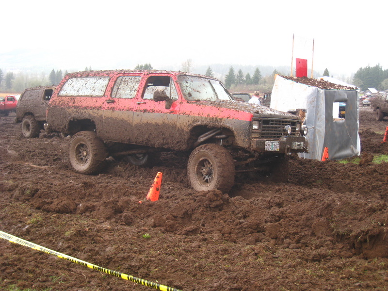 Red SUV covered in mud, driving through a mud pit with orange cones and a tent structure nearby