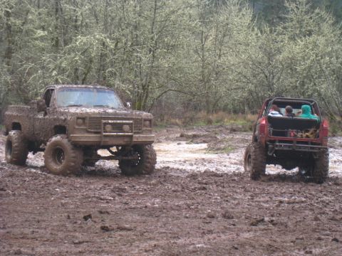 1978 Ford Bronco - Two off-road vehicles are covered in mud while navigating a wet, wooded area.