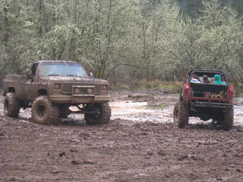 Mud-covered pickup truck and red off-road vehicle in a muddy field with trees