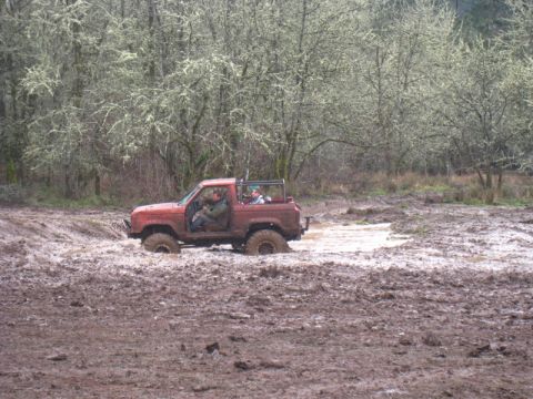 1978 Ford Bronco - A red off-road vehicle navigates through a muddy terrain, with occupants visible inside.