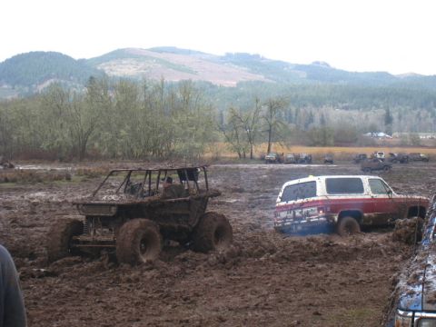 1978 Ford Bronco - Multiple off-road vehicles are stuck in a muddy field with trees and hills in the background.