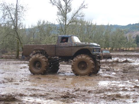 1978 Ford Bronco - Monster truck navigates a muddy terrain with trees and hills in the background.