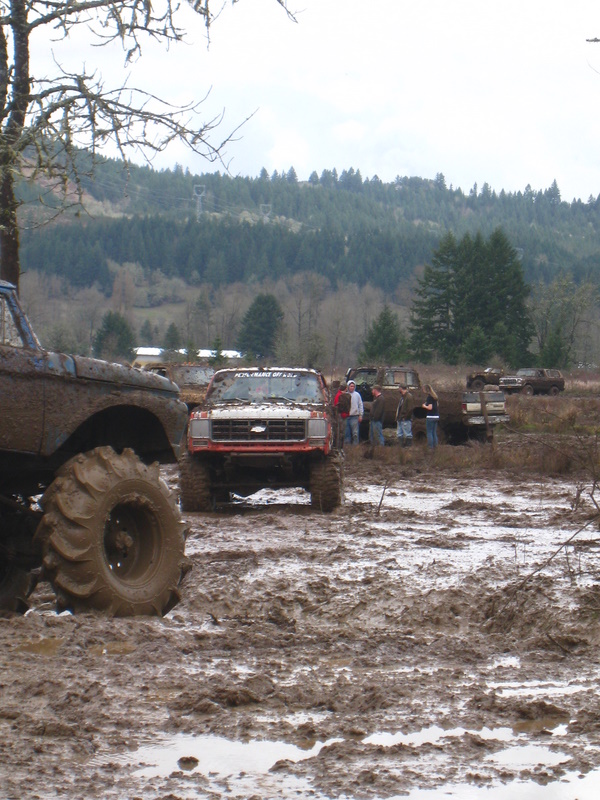 Red pickup truck with large tires in mud, front view. Other vehicles and people in background