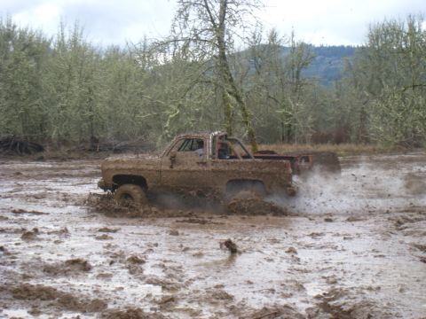 1978 Ford Bronco - A heavily muddied pickup truck churns up spray as it struggles through a muddy field with another vehicle visible behind it.