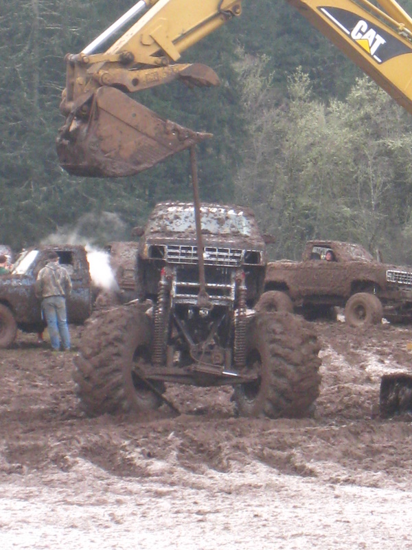 Monster trucks covered in mud, one being lifted by an excavator bucket