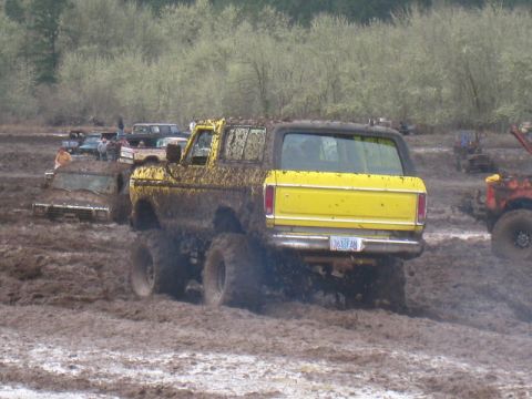 1978 Ford Bronco - A yellow SUV navigates a muddy terrain, with other vehicles visible in the background.