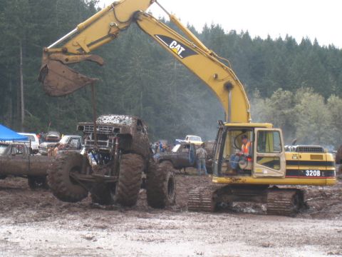 1978 Ford Bronco - Excavator lifts a mud-covered monster truck during an event in a muddy field.