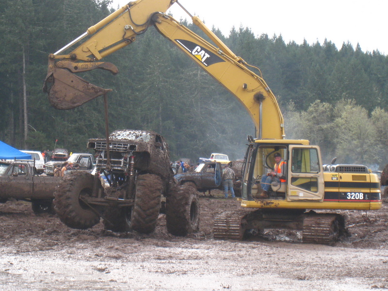 Monster truck stuck in mud being lifted by excavator; other trucks and people in background