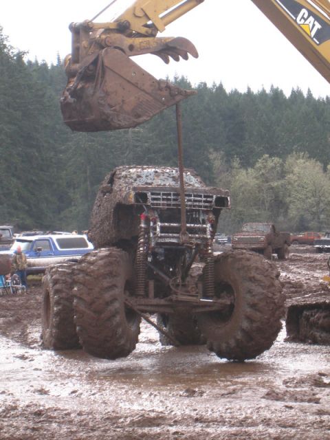 1978 Ford Bronco - An excavator is about to dump more mud onto a heavily soiled monster truck in a muddy field.