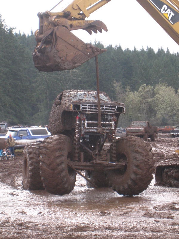 Monster truck stuck in mud, excavator bucket overhead. Other trucks in background