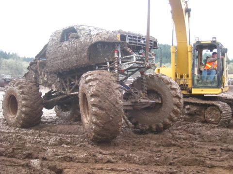 1978 Ford Bronco - A heavily mud-caked 1978 Ford Bronco monster truck is positioned next to a yellow excavator in a muddy environment.