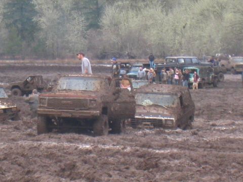 1978 Ford Bronco - Several trucks are stuck in deep mud with onlookers observing the scene.