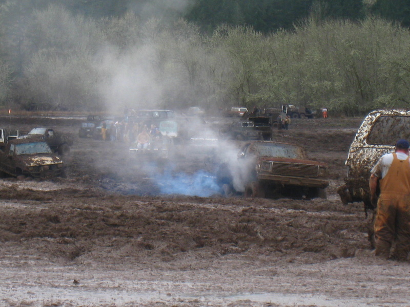Mud bogging scene with trucks stuck, one emitting blue smoke