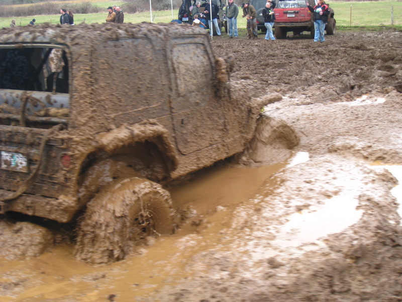 Mud-covered off-road vehicle stuck in mud pit, other vehicles and spectators in background