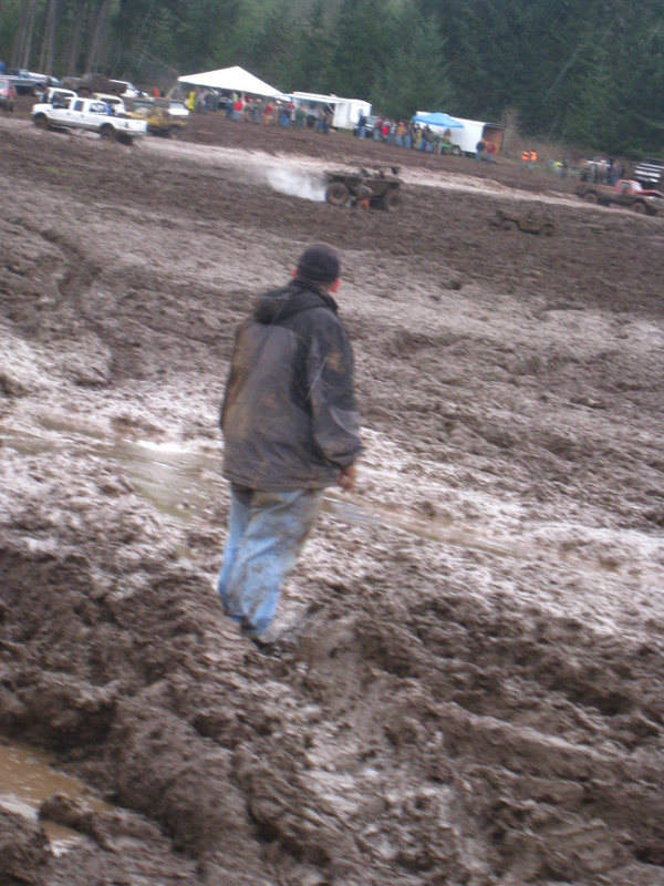 Person watching ATVs in a mud pit with tents and spectators in background
