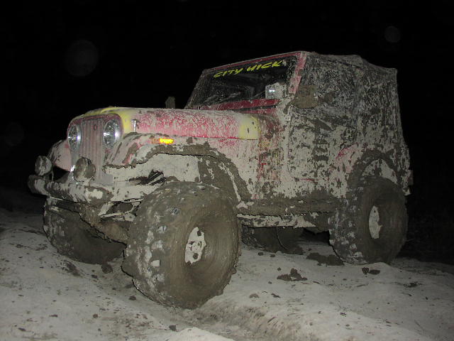 Mud-covered red Jeep CJ-7 with yellow hood, large off-road tires, and a soft top
