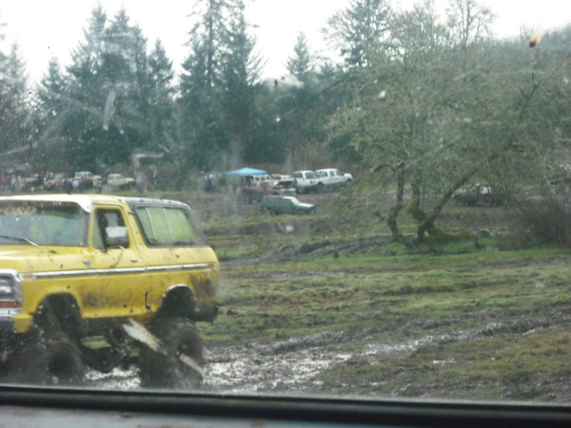 Yellow 1978 Ford Bronco with large tires splashing through mud. Other vehicles and trees in background