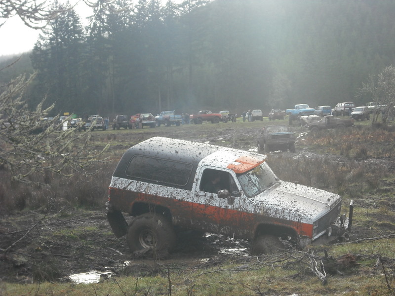 Mud-covered white and orange SUV stuck in mud, with other vehicles in background