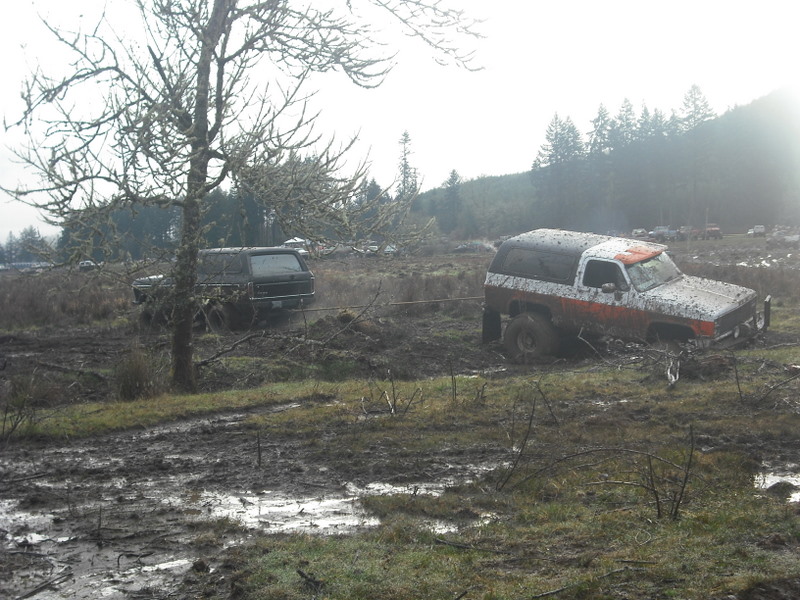 Two SUVs, one black and one white with orange stripes, stuck in a muddy field. A tow rope connects them