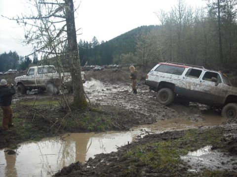 1978 Ford Bronco - Several off-road vehicles navigate a challenging muddy terrain with trees and hills in the background.