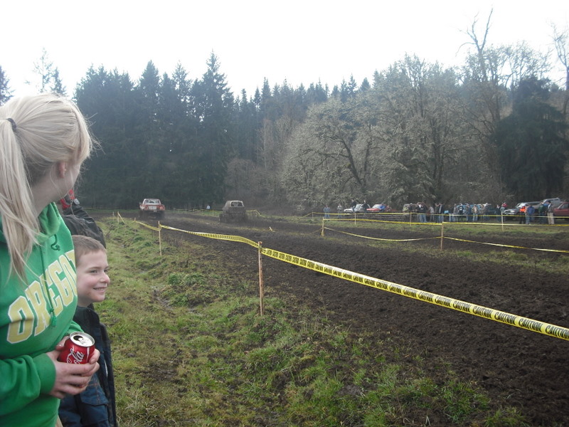 People watch vehicles race through a muddy track with trees in the background