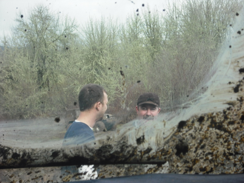 Two men look out from inside a vehicle covered in mud, with trees in the background