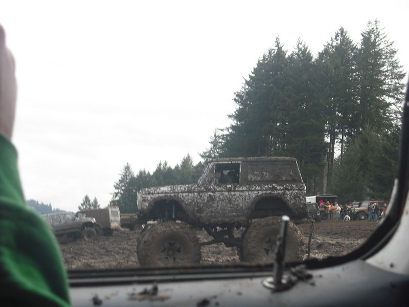 Muddy SUV with large tires driving through a muddy field with trees in the background. Other vehicles are visible in the distance