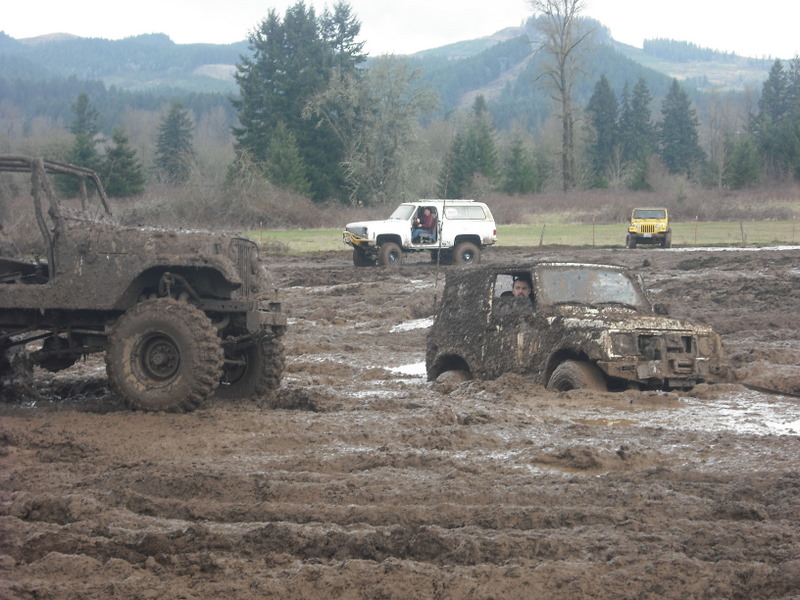 Mud-covered off-road vehicles stuck in a muddy field with trees and hills in the background