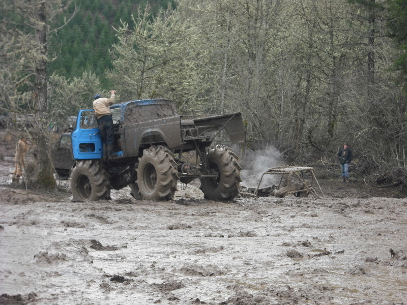 Big blue monster truck with huge tires stuck in mud, another smaller vehicle is also stuck nearby