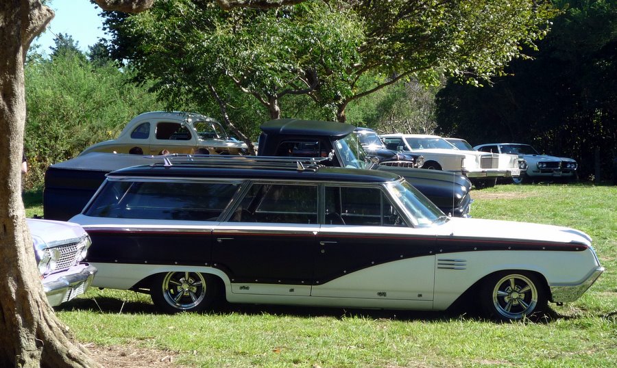 Lowered black and white 1960s station wagon with mag wheels and roof rack at a grass car show