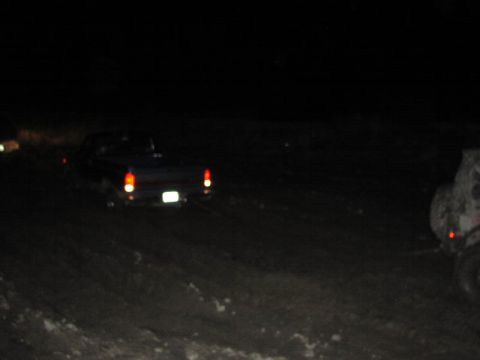 1981 Ford Bronco - A dark pickup truck is seen from the rear driving on a dirt road at night, with another vehicle partially visible to the right.