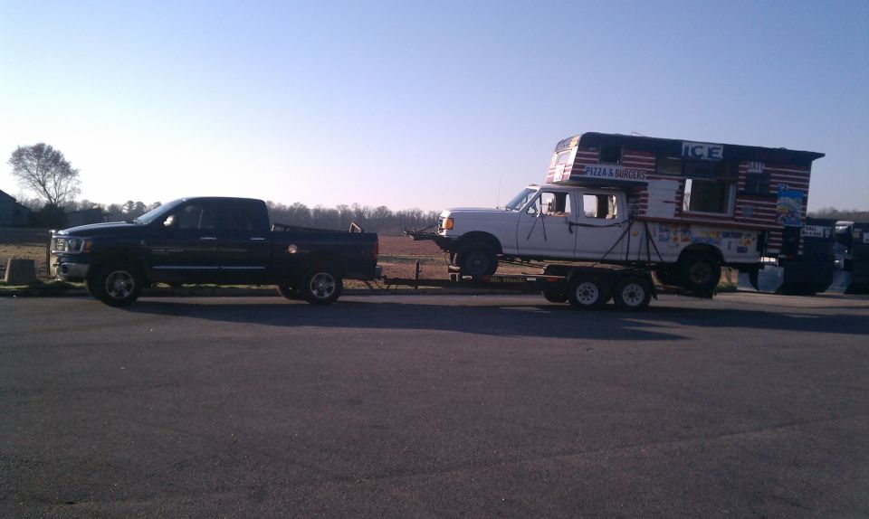 Black pickup truck towing a trailer carrying a white pickup truck with a camper shell. Camper has American flag design and 'ICE' text
