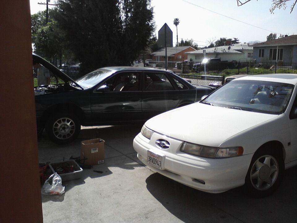 White 1996 Ford Taurus sedan parked in driveway, front quarter view. Green sedan with hood open behind it