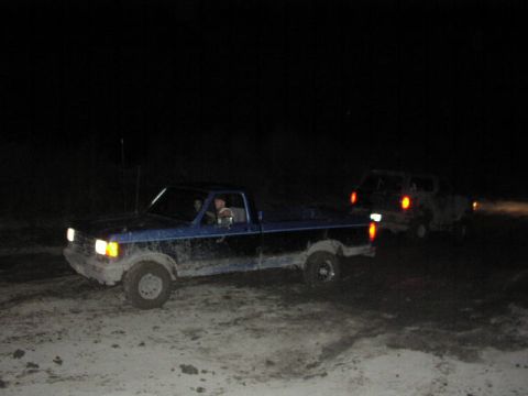 1981 Ford Bronco - Two trucks navigate a muddy terrain in the dark, with headlights illuminating the path ahead.
