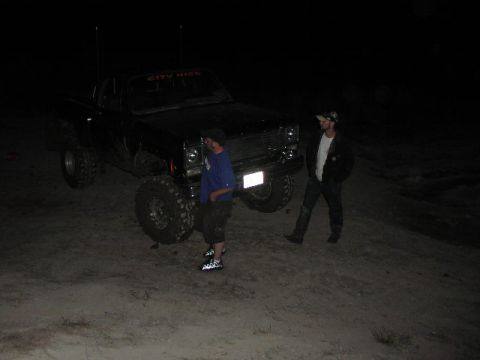 1981 Ford Bronco - A black pickup truck with large tires is parked on sandy terrain at night with two men nearby.