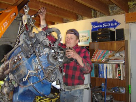 1979 Ford Mustang - Man works on a blue engine being lifted by an engine hoist in a garage.