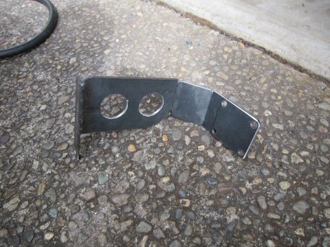2008 Jeep Wrangler - A fabricated black sheet-metal bracket lying on an exposed-aggregate concrete driveway, with two round lightening/pass-through holes in the…