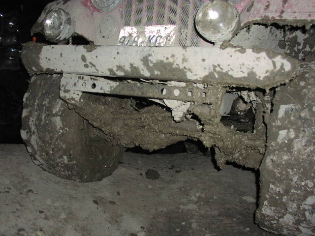Mud-covered front bumper, tire, and grille of a vehicle. License plate visible