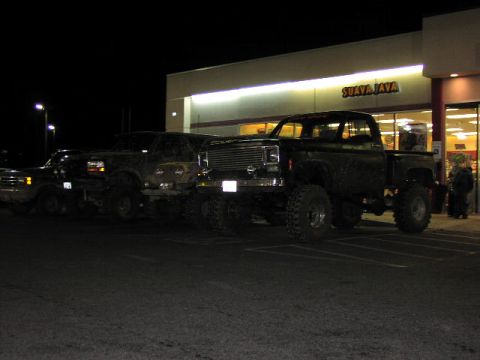 1981 Ford Bronco - Multiple heavily modified pickup trucks are parked outside a "SUAVA JAVA" establishment at night.