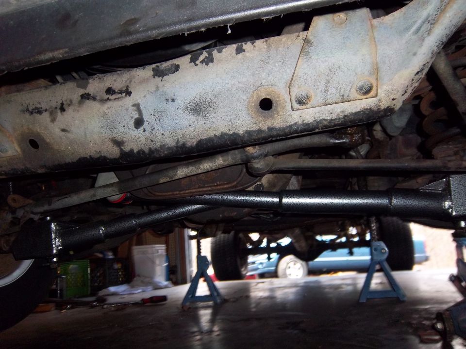 Underside of a vehicle on jack stands, showing rusty frame and new black steering components. Another vehicle is visible in the background